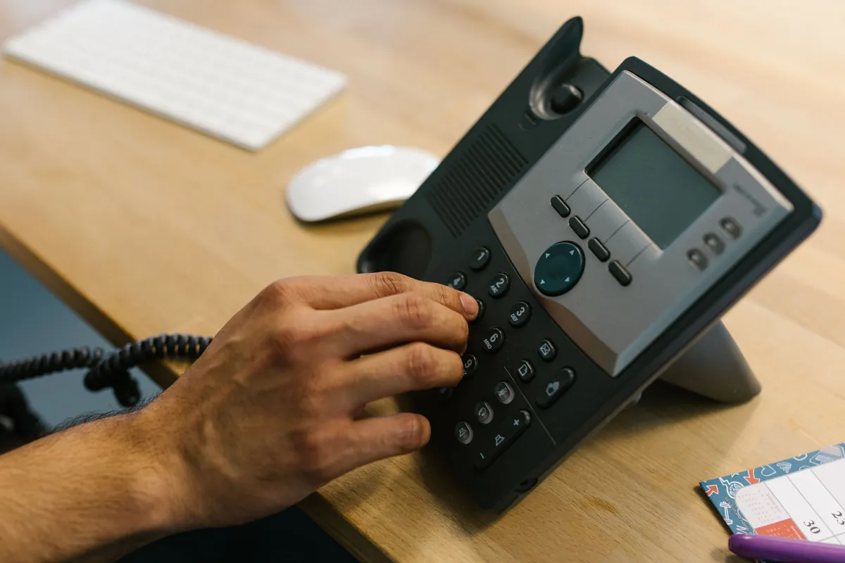 A hand dialing an office desk phone beside a keyboard and a wall planner
